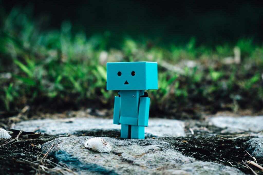 Cute blue toy robot standing on a rocky path surrounded by greenery.