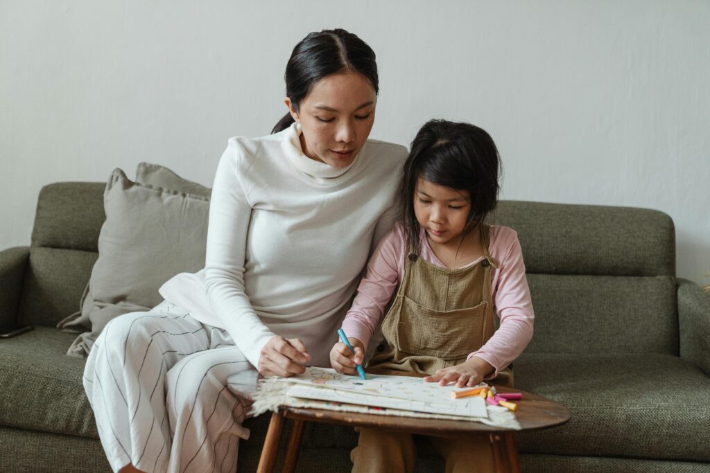Concentrated little Asian girl in casual clothes studiously drawing picture on paper while mother watching process sitting together on sofa
